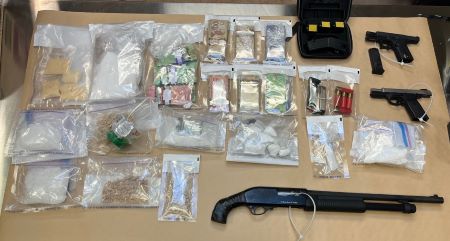 Evidence bags containing cash, suspected drugs, and ammunition, are displayed on a table along with two handguns and long gun. 