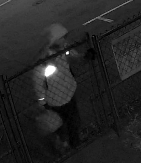 Une image en noir et blanc d&rsquo;une personne debout devant une cl&ocirc;ture &agrave; mailles m&eacute;talliques, tenant un jerrycan dans sa main droite.