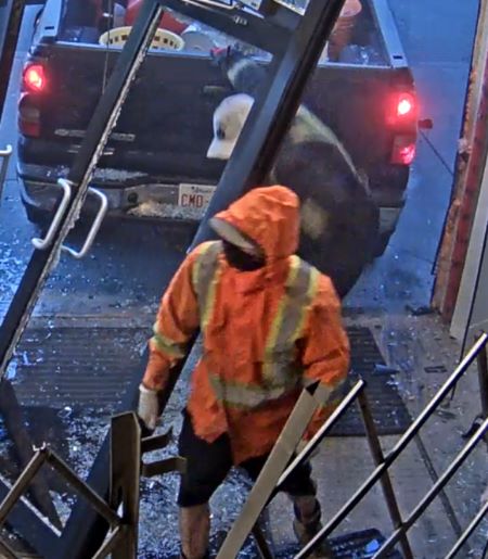 Un suspect v&ecirc;tu d&rsquo;une veste &agrave; haute visibilit&eacute; orange et jaune avec la capuche relev&eacute;e, se tenant devant le suspect en combinaison bleue. Les feux arri&egrave;re d&rsquo;une camionnette de couleur fonc&eacute;e s&rsquo;allument derri&egrave;re eux. 