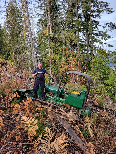 Police officer stands atop a recovered stolen green and yellow bulldozer surrounded by dense brush.