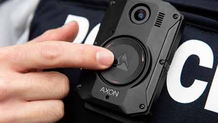 Photo of a police officer wearing the body worn camera, his finger is pointing to the camera on his police vest.