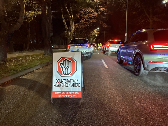 pop-up sign on the road that reads <q>CounterAttack road check ahead, have your driver&rsquo;s licence ready</q> with a police vehicle and cars in the background