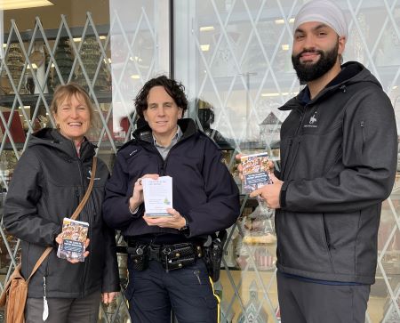 Two volunteers and a uniformed police officer stand in front of a display window with crime prevention tip cards in their hands. 
