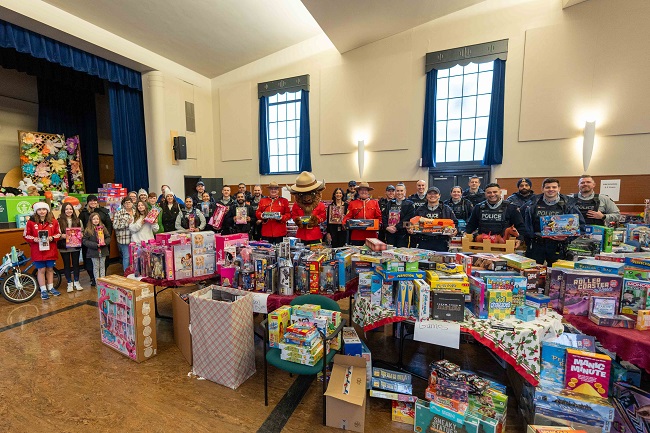 Two RCMP officers in red serge with RCMP mascot Sgt. Bison. A group photo of RCMP officer in uniform standing behind a large pile of toys.  RCMP mascot Sgt. Bison giving a hih five to a small child.  Group photo of Blundell Family with RCMP officers. Photo of Christmas tree with toys.