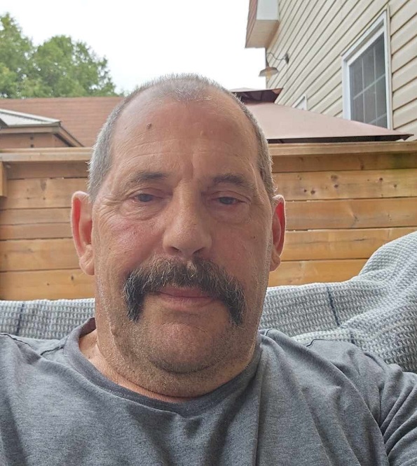 Photo of a caucasian male with a dark grey moustache, blue eyes and a balding buzz cut of brown hair. Male is wearing a grey t-shirt.