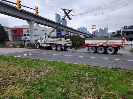 A side view of a dump truck that had collided with a hydro pole and a lamp post on the side of the road, just before a train track and the SkyTrain.
