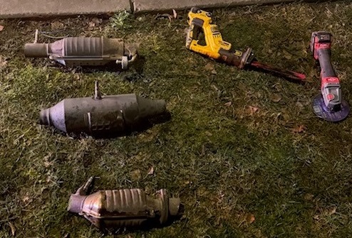 Three catalytic converters laying on the grass on the side of the road, beside a red and black grinder and a yellow reciprocal saw.