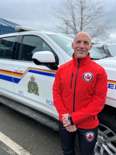 Cst. Paul Jones, in red a Wounded Warrior jacket and black Wounded Warrior pants standing in front of a police vehicle in the parking lot of the Comox Valley RCMP Detachment