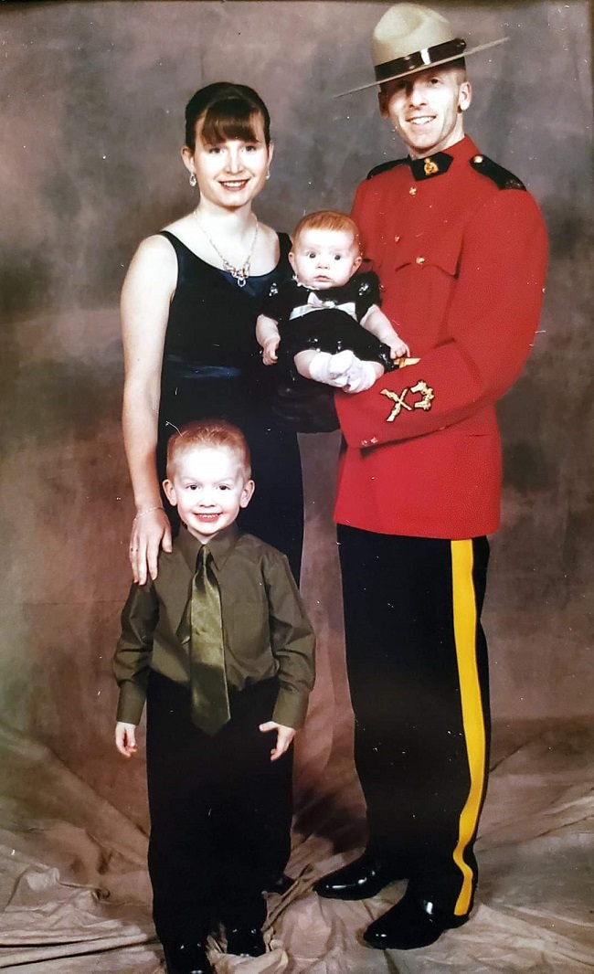 Photo of Cpl. Scotty Schumann, wearing his RCMP red serge uniform at his graduation from RCMP Training Academy. The photograph includes his wife standing beside him wearing a black dress along with his son, age 4 wearing a green shirt with a tie and black pants. Cpl. Schumann is holding his 4-month-old daughter in his arms.