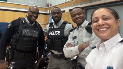 Cst. Dwight Martin stands smiling beside three other police officers in a school gym during a Black Excellence Day event at Cloverdale Traditional School (Left to right: Cst. Dwight Martin, Cpl. George Amoako, Cst. Berthier Kyobela, and Insp. Veronica Fox)