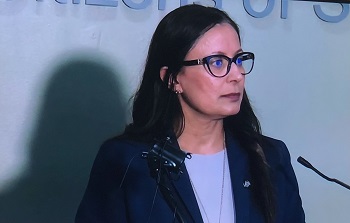 Cpl. Dhesi with wearing a blazer and glasses speaking to the media. 