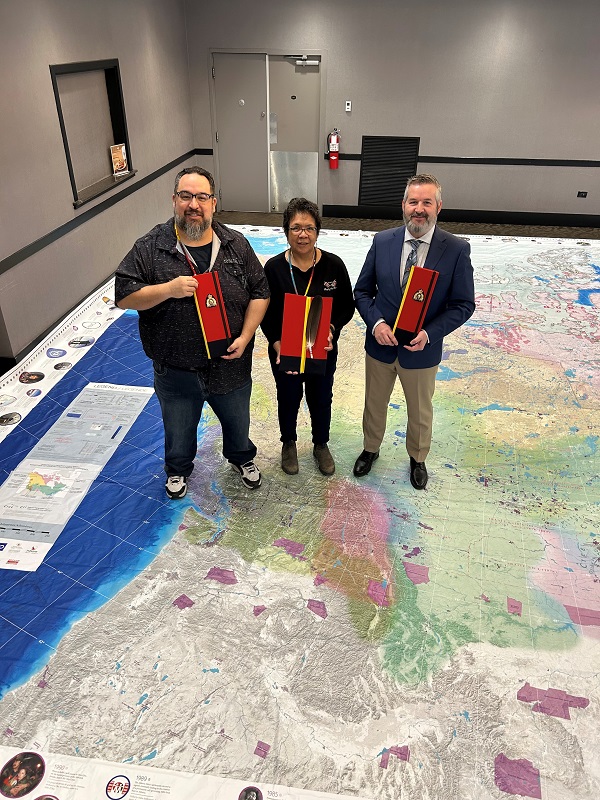 From left to right are Members of the BC Indigenous Cultural Advisory committee Jason Campbell of Seabird Island First Nation, Tina Donald of Simpcw First Nation, and Acting Officer in Charge of Indigenous Policing Services, Staff Sergeant Steve Pebernat, each holding an eagle feather in a specialized RCMP storage case, standing together on a large map of Turtle Island (North America).