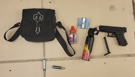 A black satchel, a five-dollar bill, a canister of bear spray and a black pellet hand gun laying on a tile yellow floor.