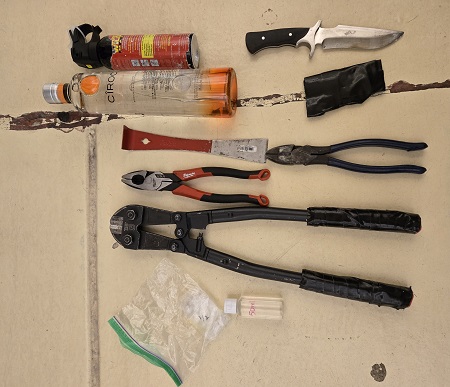 Various tools, including pliers, a bolt cutter, a snipping tool, a small pry bar, a black and silver hunting knife, a red canister of bear spray and a bottle of liquor on a yellow tile floor.