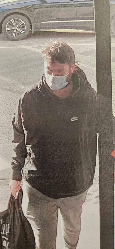 The suspect walks through a doorway wearing a dark Nike jacket and beige pants, while holding a black bag with white writing.