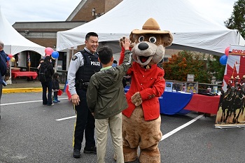Safety Bear, wearing a red serge standing next to a male Mountie. Safety Bear gives high five to a member of the public. 