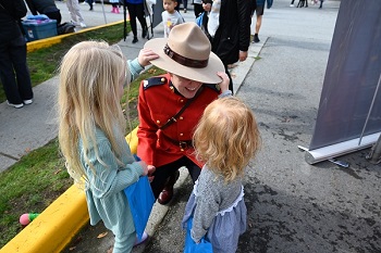 A Mountie in red serge kneeing down while two little girl&rsquo;s play with the mountie&rsquo;s Stetson 