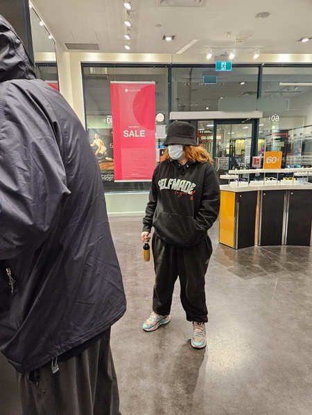 Photo of female suspect wearing a medical mask, black hat, black sweatshirt, black sweatpants and white runners with blue stripes and holding a cannister in her right hand.