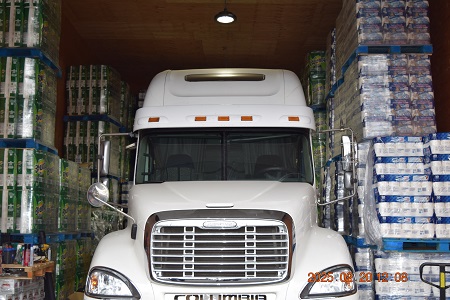 A white semi tractor is parked in a warehouse with pallets of toilet paper and paper towels stacked behind and beside it.