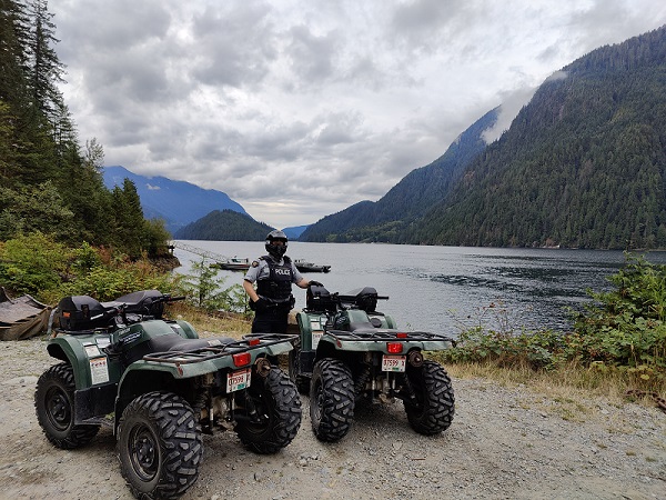 A photo of an RCMP Member wearing a full-face helmet, standing in front of two ATVs with a view of forested area, a lake and mountains in the background