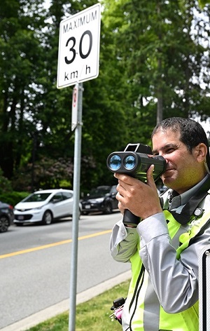 A male RCMP Officer using a laser speed gun, next to a 30km/h speed sign
