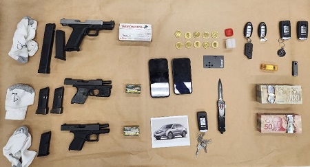 Seized evidence laid out on a table, including three hand, five magazines, two cellular phones, five vehicle fobs, a switch blade and two bundles of Canadian cash.