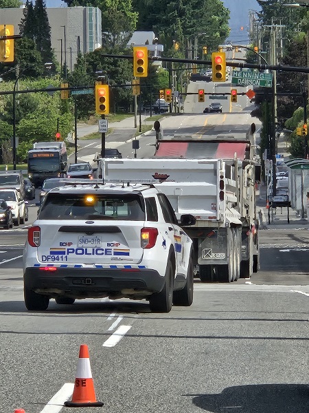 An RCMP BC Highway patrol police vehicle is driving being a large full dump truck on a busy street in Burnaby.