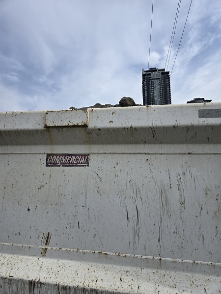 The side of a dump truck with some of the load inside, including some rocks, parked on the side of the road with a high-rise residential building in the background.