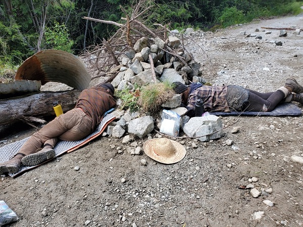 Photo of two individuals on the roadway, surrounded by rocks, with their arms attached to locking devices in the ground