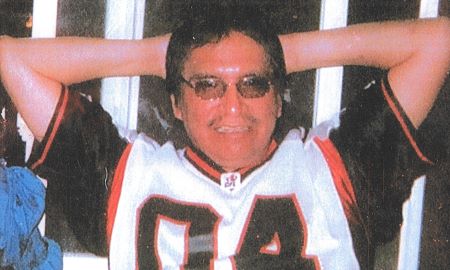 Fred Yellow Old Woman sits with his arms behind his head. He is wearing a black, white, and red, with a number four on it, as well as dark glasses.