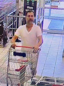third male suspect pushing a cart into the store. He is wearing a white v-neck tshirt and light coloured shorts. He has short dark hair and a greying beard or 5 o&rsquo;clock shadow. 