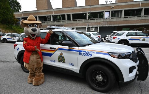 A bear mascot wearing a Red Serge poses outdoors beside a Burnaby RCMP SUV beside a brick building
