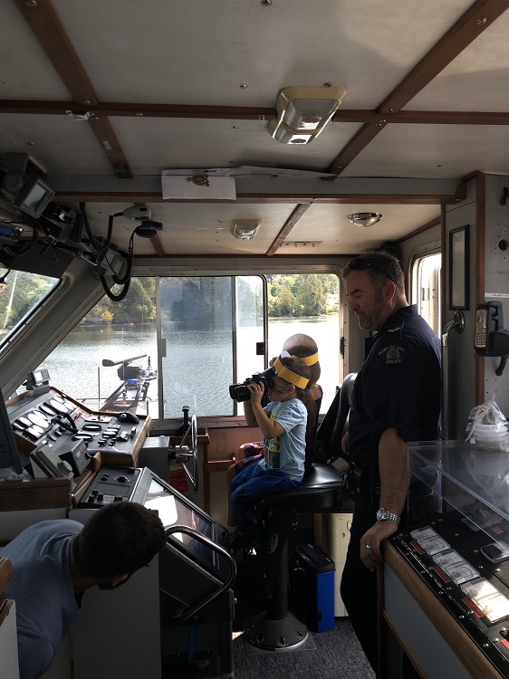 An RCMP officer and a young boy on the bridge of a police ship. The boy is wearing a police-style hat and looking through binoculars