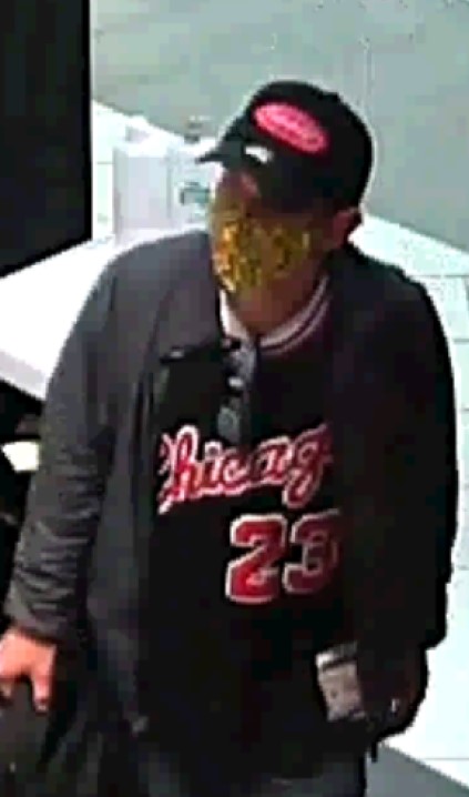 A man standing wearing a dark baseball hat with a red logo, a grey jacket and a dark shirt with the word Chicago and number 23 on the front of it, carrying a black back pack in his right hand.