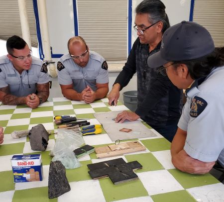 D&eacute;monstration : Des policiers en uniforme sont rassembl&eacute;s autour d&rsquo;Ed Jensen pendant qu&rsquo;il fait une d&eacute;monstration de technique de taille de silex sur une table &agrave; carreaux verts et blancs. Des bandages, des outils et d&rsquo;autres fournitures sont pos&eacute;s sur la table. 