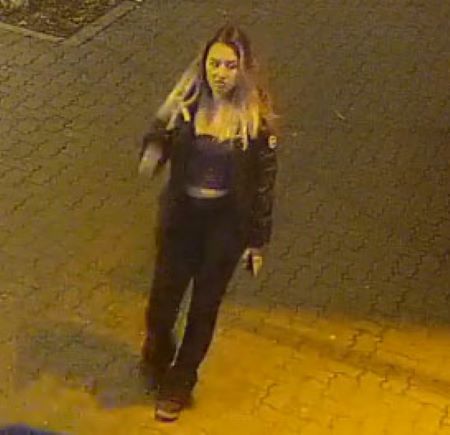 Photo of a woman who accompanied the man. In this image, Her long hair is down and her dark coat is open. She is wearing a short sweater and dark pants. Her coat has a white logo patch stuck on its left shoulder.