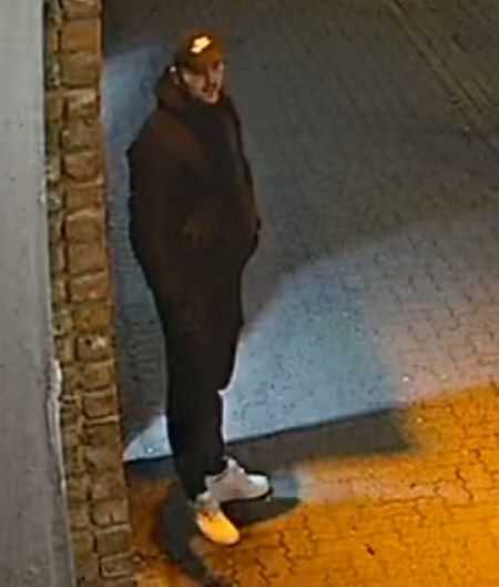 Un homme de grande taille aux poils faciaux fonc&eacute;s, qui portait une casquette fonc&eacute;e avec un logo blanc, un manteau d&rsquo;hiver et des pantalons fonc&eacute;s et des souliers blancs. 