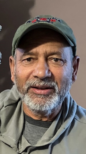 Photo of Narinder Sandhu, South Asian man with a white and grey moustache and beard wearing a green sweater and green baseball cap.