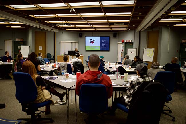 Members of the BC RCMP from various sworn and civilian ranks sitting at tables viewing slideshow on TV