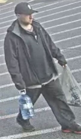 Un homme de race blanche marchant dans un stationnement, portant une casquette de baseball noire, un chandail fonc&eacute;, une veste fonc&eacute;e, un jean et des chaussures noires.